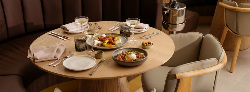 A round timber table set for breakfast in a modern restaurant, featuring plated dishes, pastries, fresh fruit, and glasses of prosecco, surrounded by upholstered seating in a warm, contemporary setting.